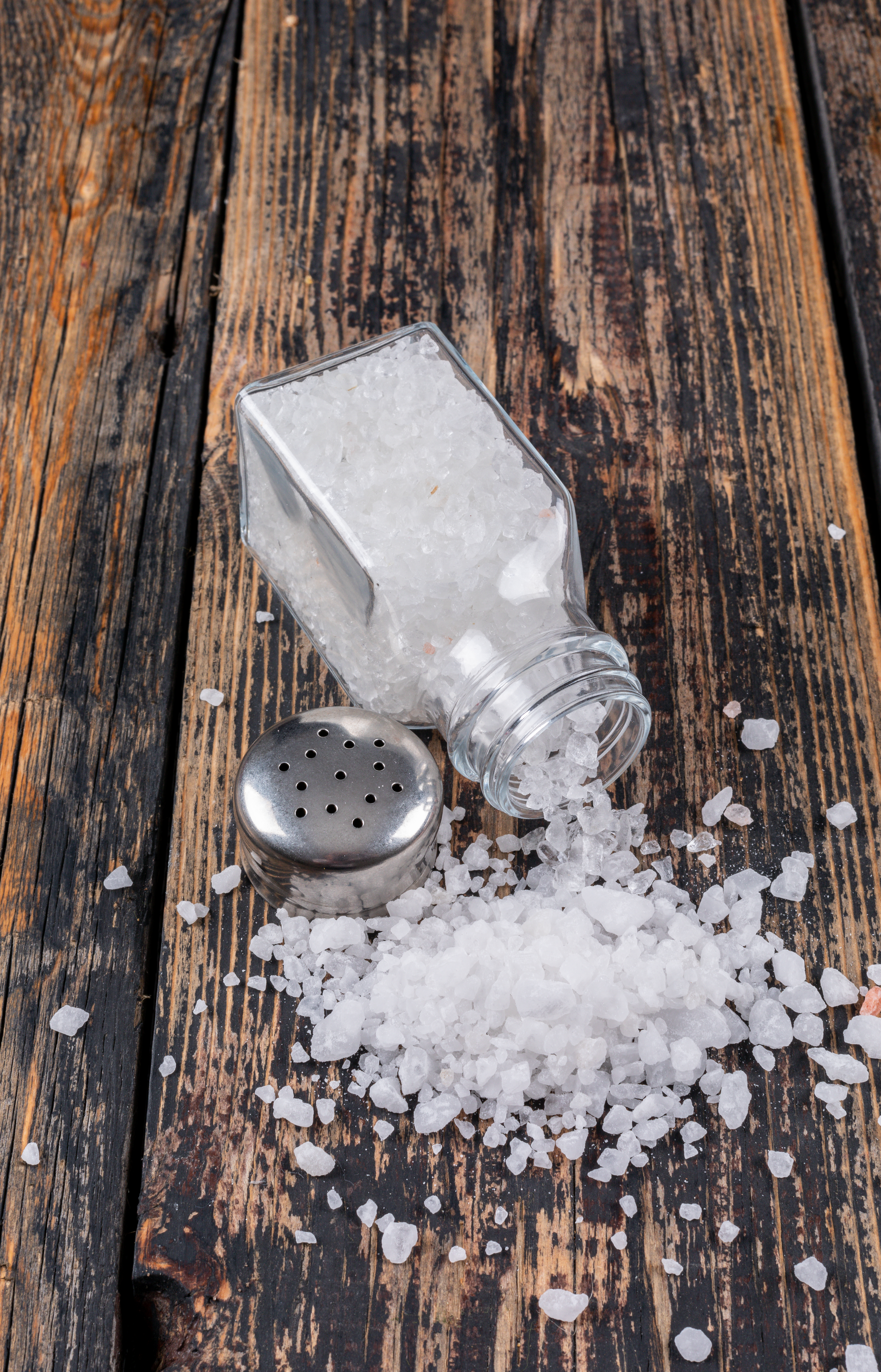 High angle view sea salt coming out of open salt shaker with its cap on dark wooden background. vertical