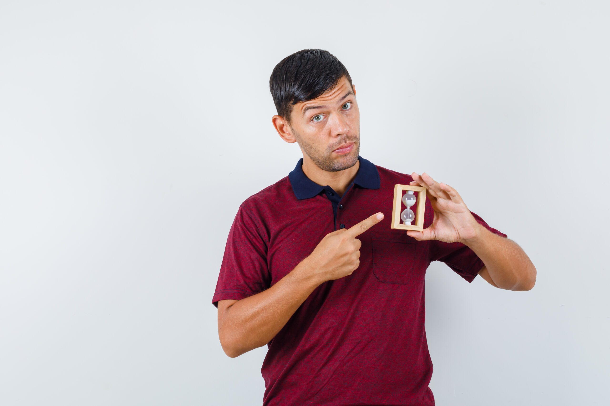 Young man pointing at hourglass in t-shirt and looking careful. front view.